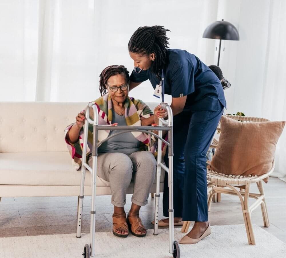 A nurse assists an elderly woman who is using a walker to stand up from a couch, both in a comfortable, well-lit room. - Home Instead