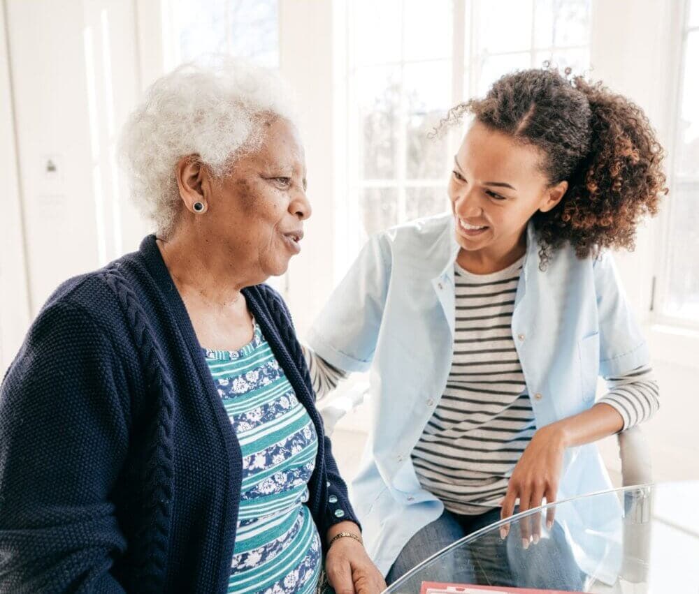 Elderly woman and young woman smiling and conversing at a table in a bright room. - Home Instead