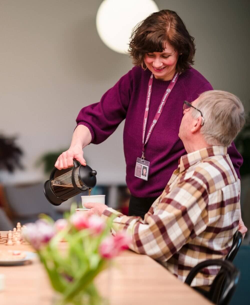 A Southbourne Care Professional in a purple sweater serves coffee to an older man seated at a table with a chessboard and flowers. - Home Instead Bournemouth & Christchurch