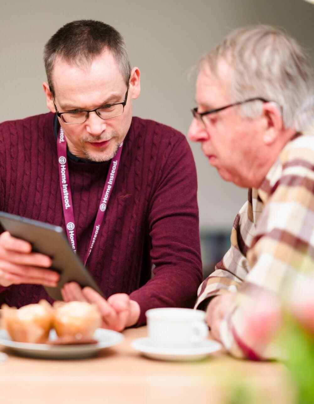 Two men looking at a tablet screen, sitting at a table with coffee cups and muffins. - Home Instead