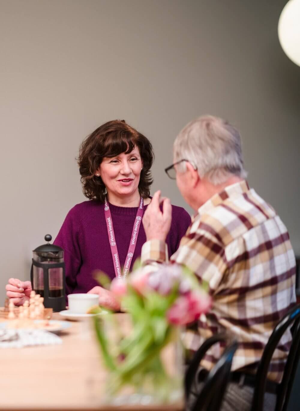 A woman pours tea for an elderly man sitting at a table, both smiling warmly in a cozy room. - Home Instead