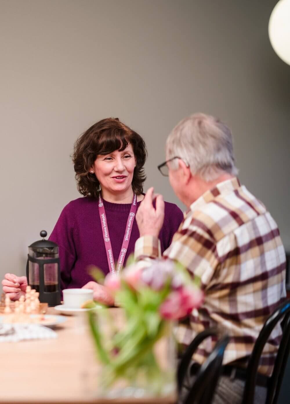 Two people having a conversation at a table with coffee and a chess set, with flowers in the foreground. - Home Instead