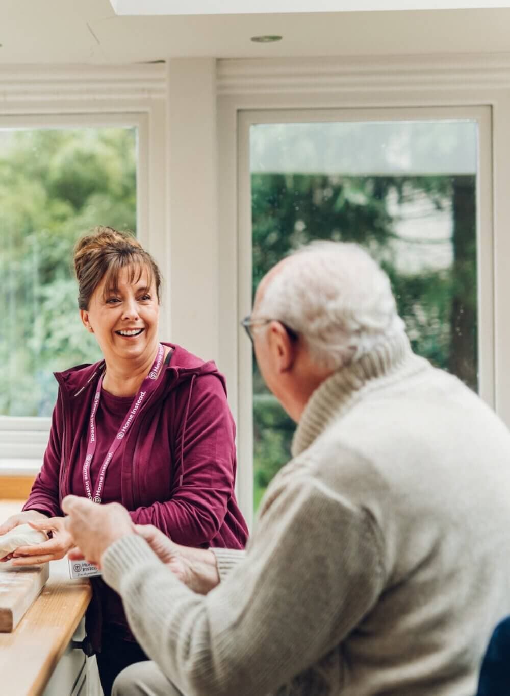 A woman with a name tag sitting at a table, smiling and talking to an older man in a gray sweater in a bright room. - Home Instead