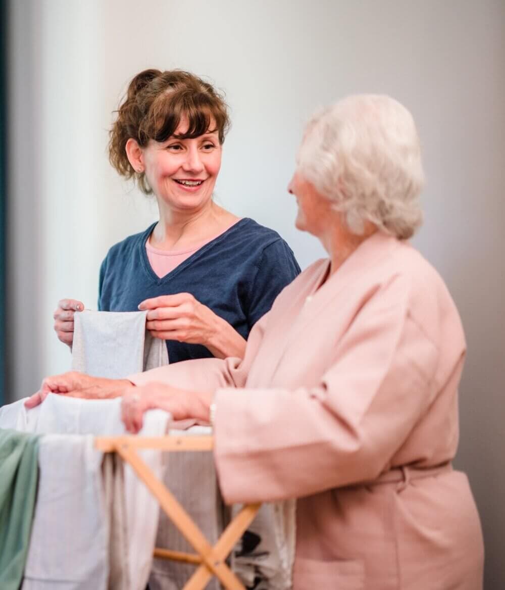 A young woman and an older woman smile while folding laundry together indoors. - Home Instead