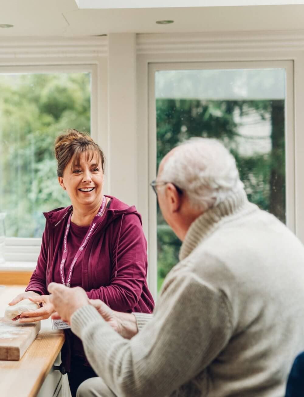 Woman in purple hoodie smiling and talking with an elderly man in a white sweater at a bright, windowed room. - Home Instead