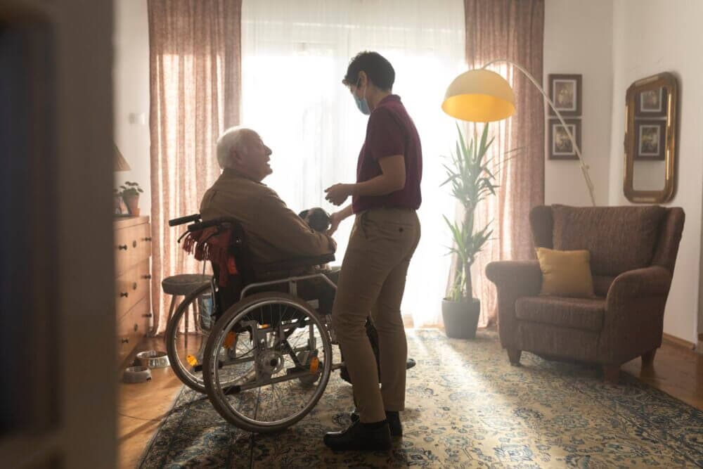 Person in a wheelchair smiling and talking to a standing caregiver in a warmly lit living room. - Home Instead