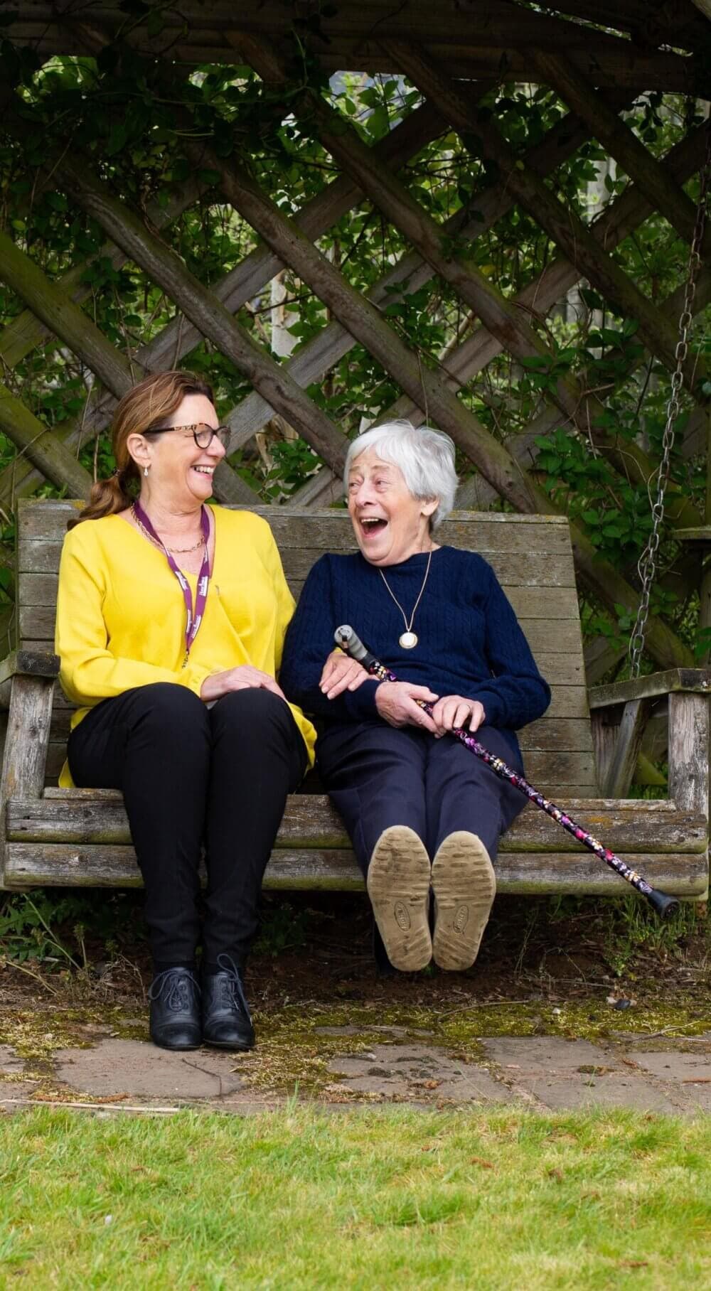 Two women sitting on a garden bench, laughing together. One has a walking stick with them. - Home Instead