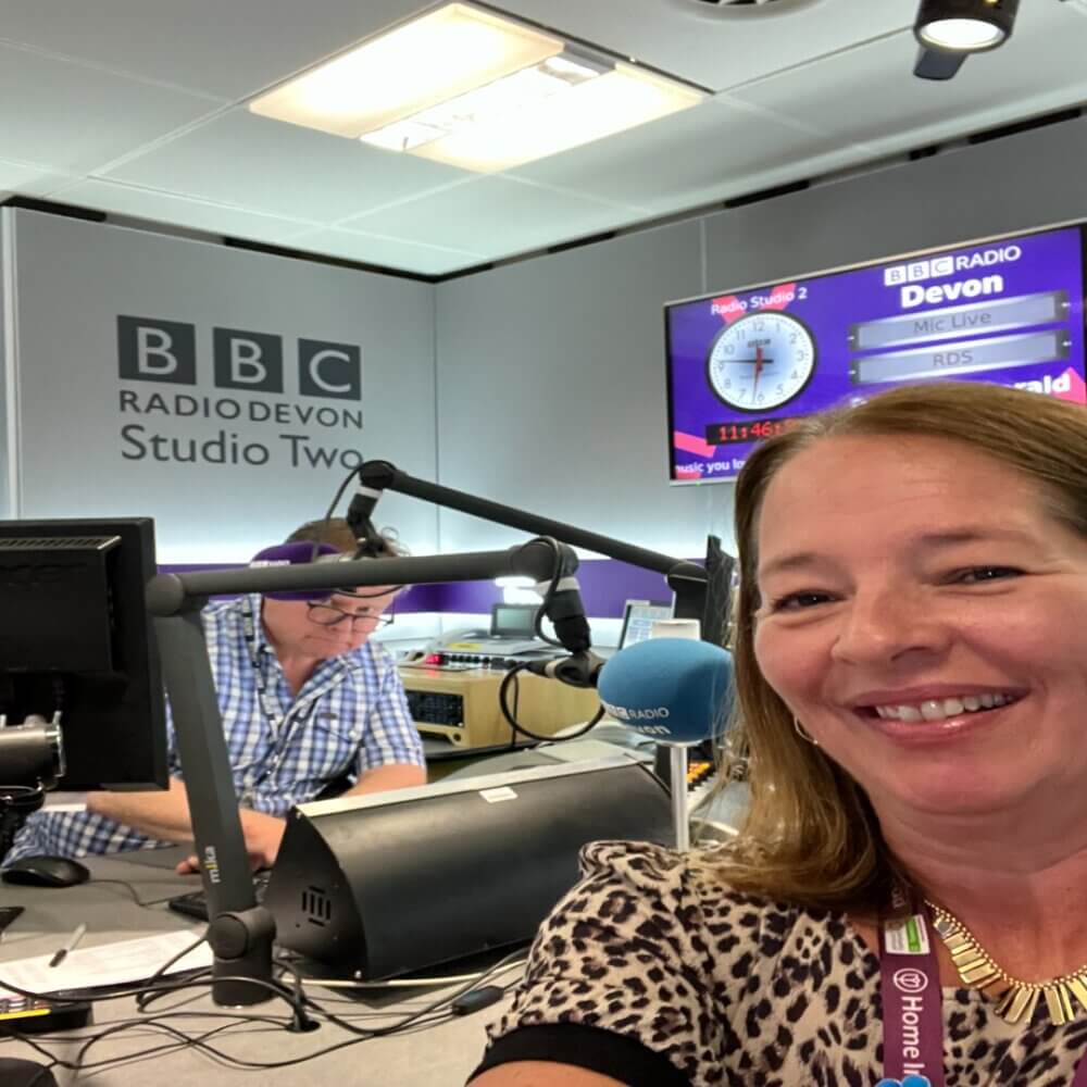 Two people in a BBC Radio Devon studio, with a woman smiling in the foreground and a man working in the background. - Home Instead