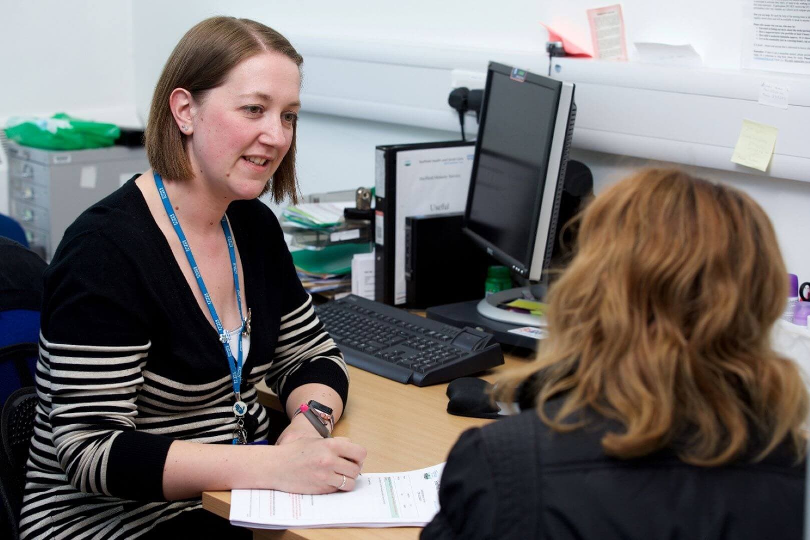 A woman wearing a striped sweater talks to another woman at a desk with a computer and papers. - Home Instead