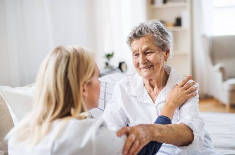 A younger woman comforts an elderly woman sitting on a couch, both smiling and holding hands in a light-filled room. - Home Instead