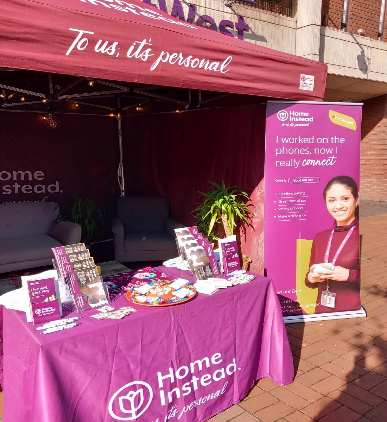 Outdoor booth with "Home Instead" branding, informational pamphlets on a table, and a banner with a smiling woman. - Home Instead