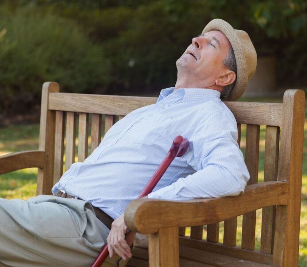 An elderly man in a hat is sleeping on a wooden bench while holding a cane on a sunny day. - Home Instead