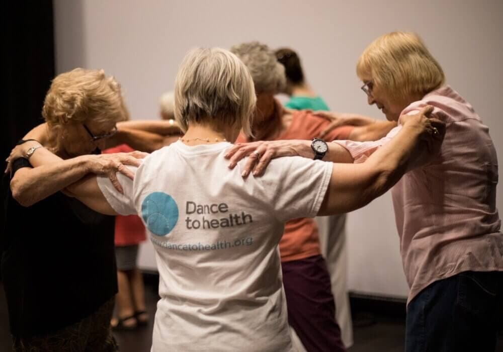 Group of elderly women dancing together, one wearing a "Dance to Health" shirt. - Home Instead