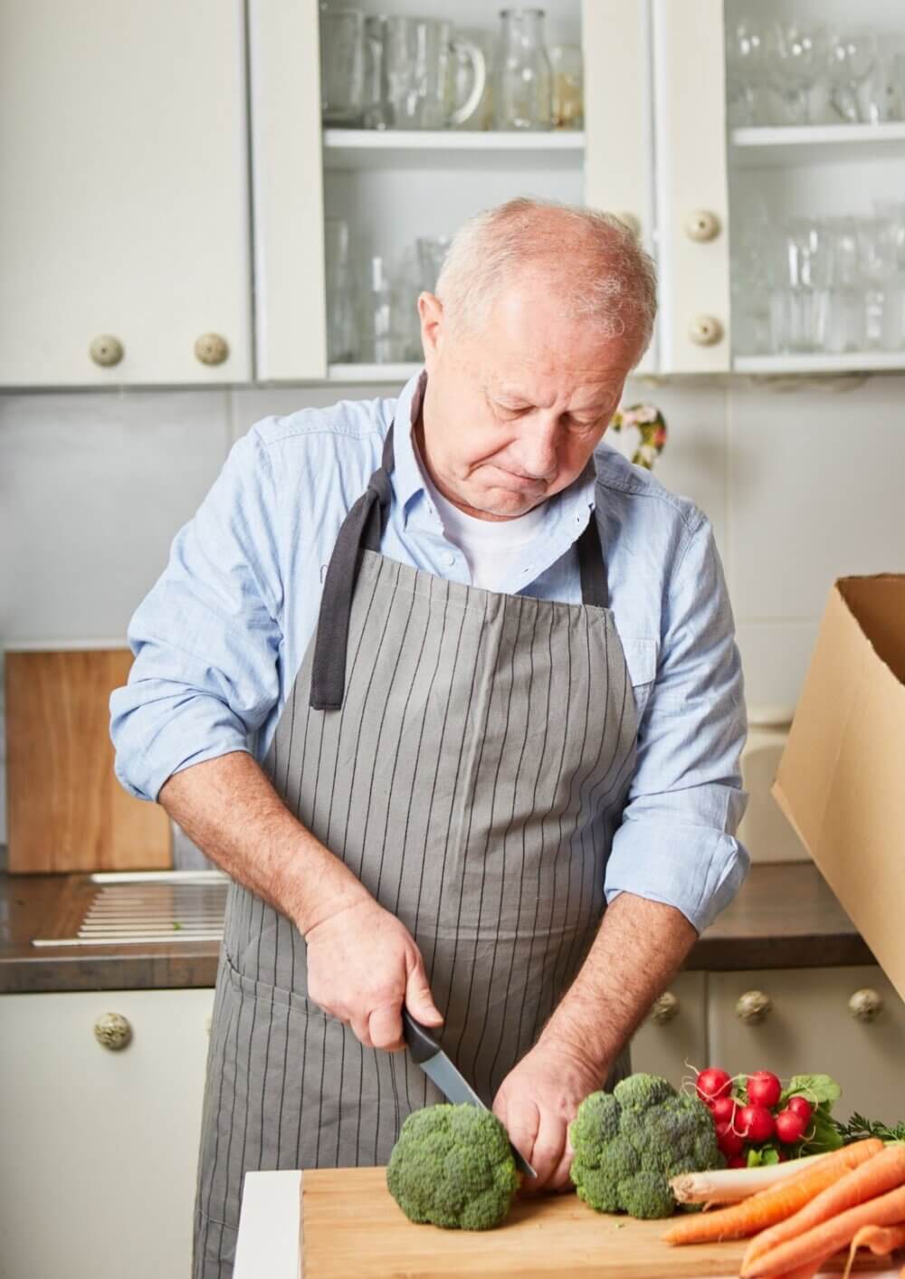 Older man in a kitchen cutting broccoli on a wooden board, wearing a gray apron and a blue shirt. - Home Instead