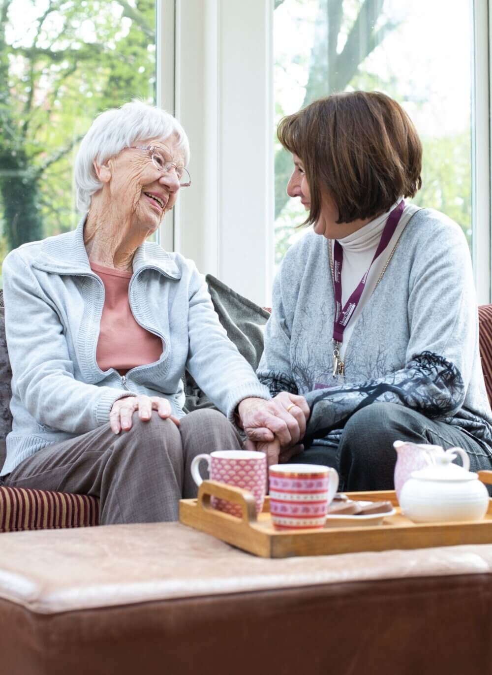 Two women, one younger and one older, smiling and playing video games on a gray sofa in a bright living room. - Home Instead
