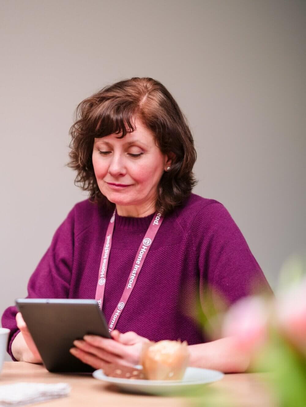 Woman in a purple sweater looking at a tablet, wearing a lanyard, with a muffin and a cup on the table. - Home Instead