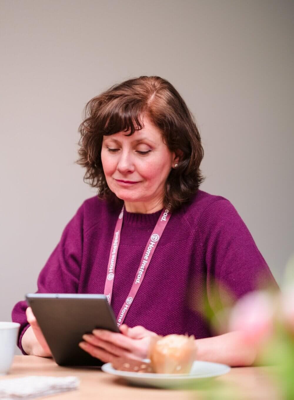 Woman in a purple sweater reading a tablet while sitting at a table with a muffin and a cup. - Home Instead