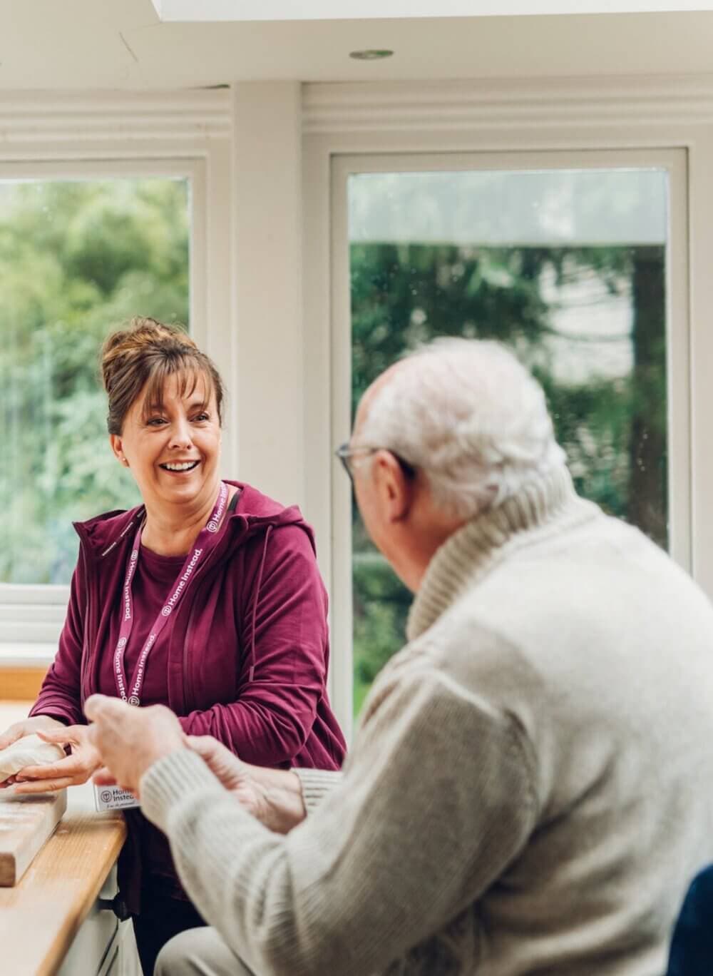 A woman smiling and talking with an elderly man in a bright room with large windows. - Home Instead
