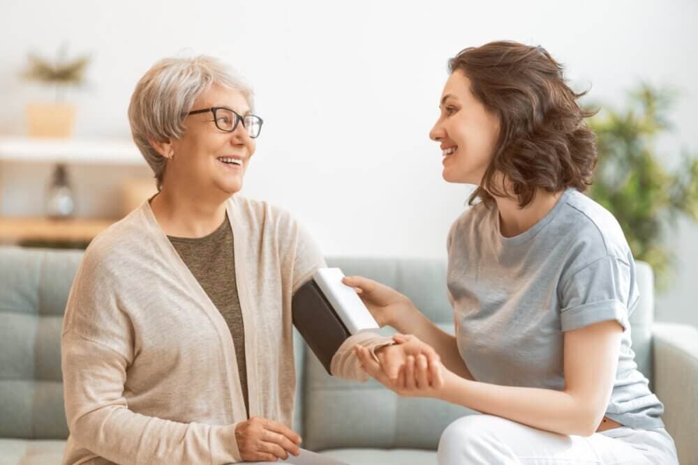 A smiling young woman takes the blood pressure of a seated older woman using a blood pressure monitor. - Home Instead