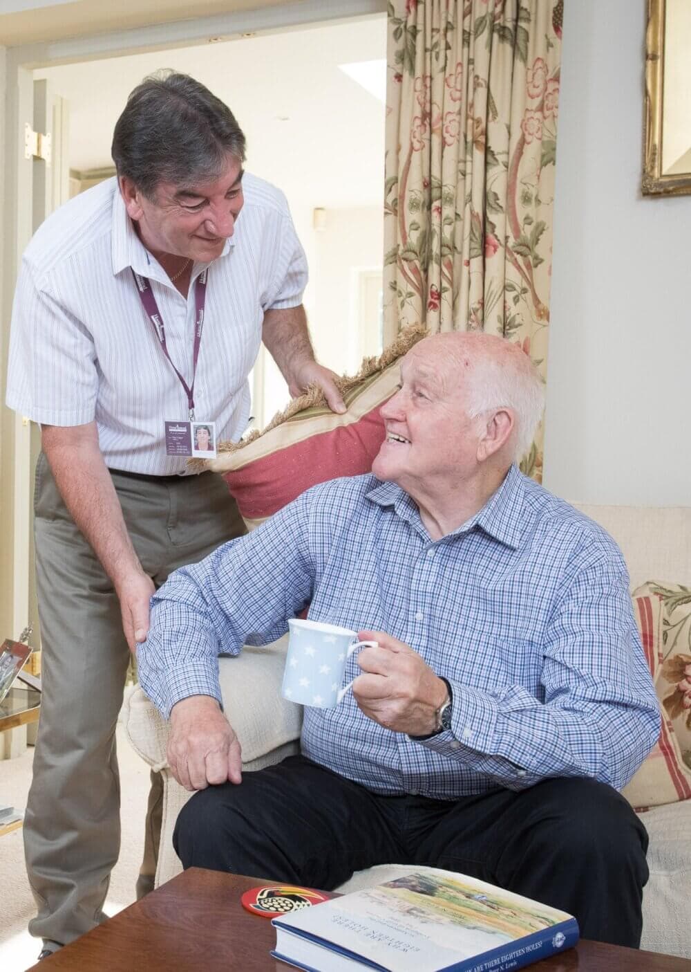 Two elderly men, one sitting and holding a mug, the other standing and smiling, in a warmly decorated living room. - Home Instead