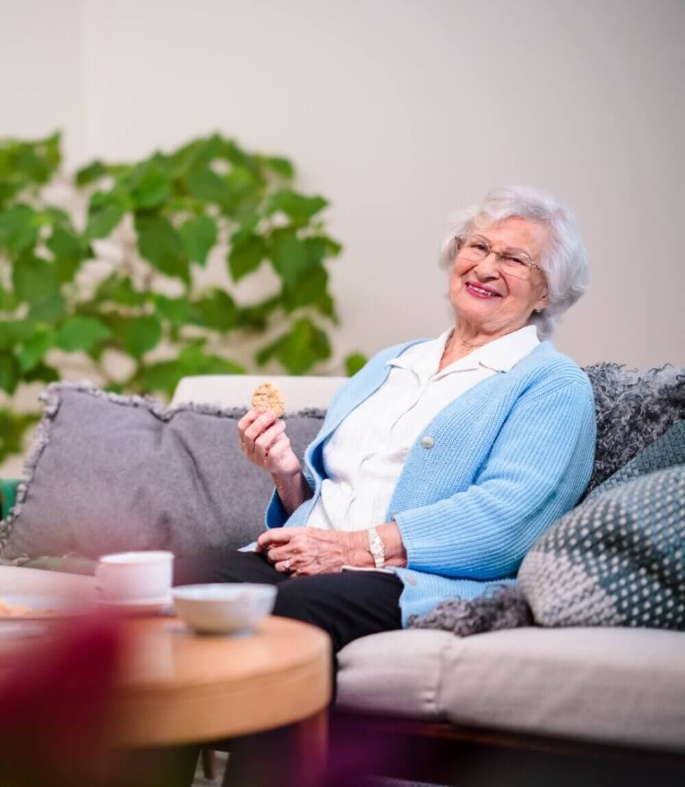 Elderly woman in a blue cardigan sitting on a couch, smiling and holding a cookie. - Home Instead