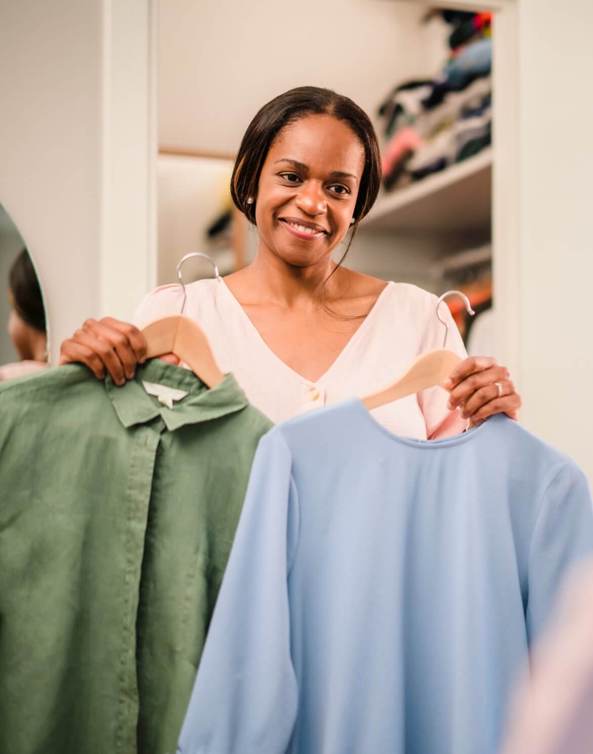 Woman smiling and holding up a green shirt and a blue shirt on hangers in front of a wardrobe. - Home Instead
