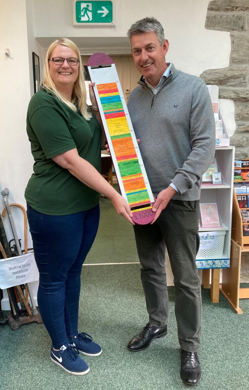Two people smiling and holding a colorful fundraising thermometer chart in a room with stone walls and bookshelves. - Home Instead