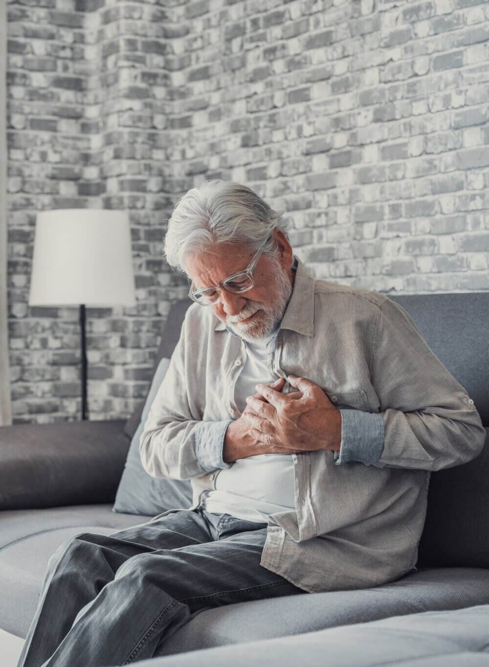 Elderly man sitting on couch, clutching chest in apparent pain, with a worried expression on his face. - Home Instead