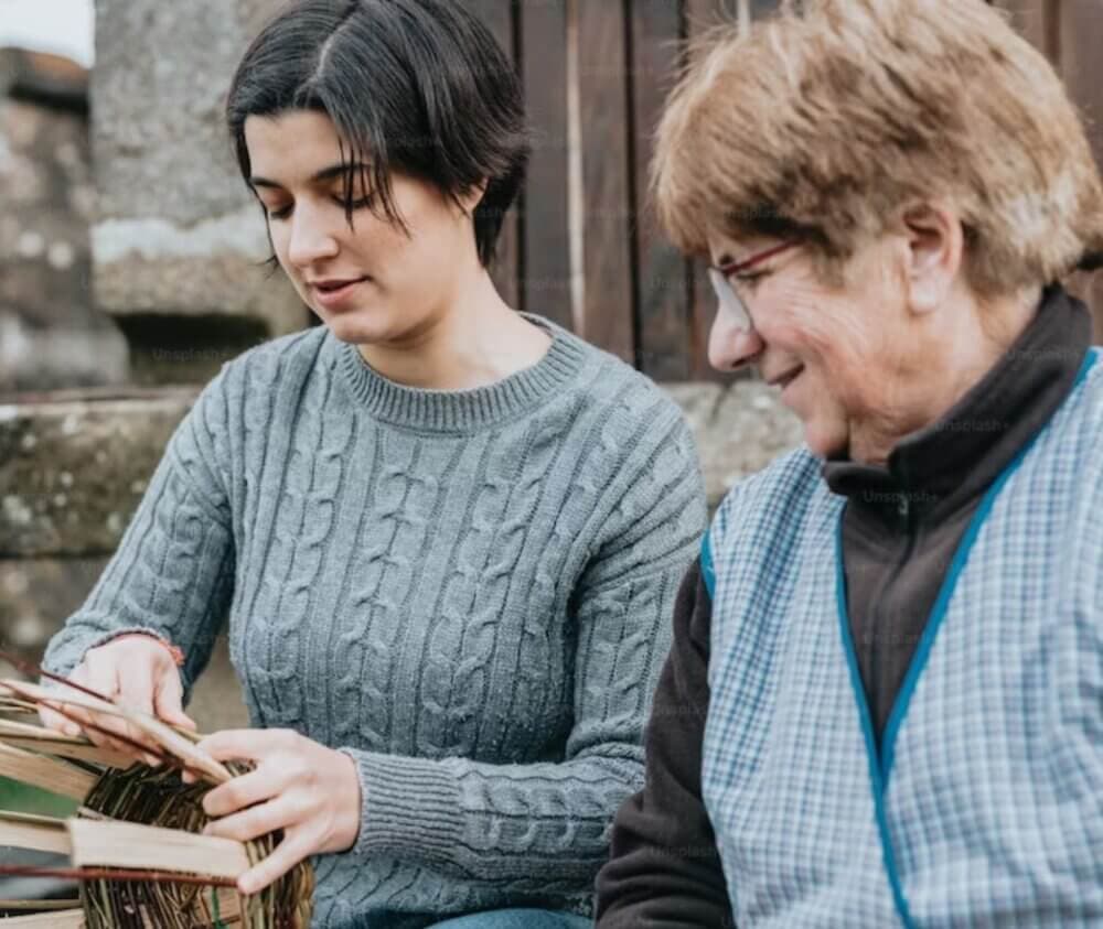 Two women sit outdoors, one weaving a basket while the other smiles and watches. - Home Instead