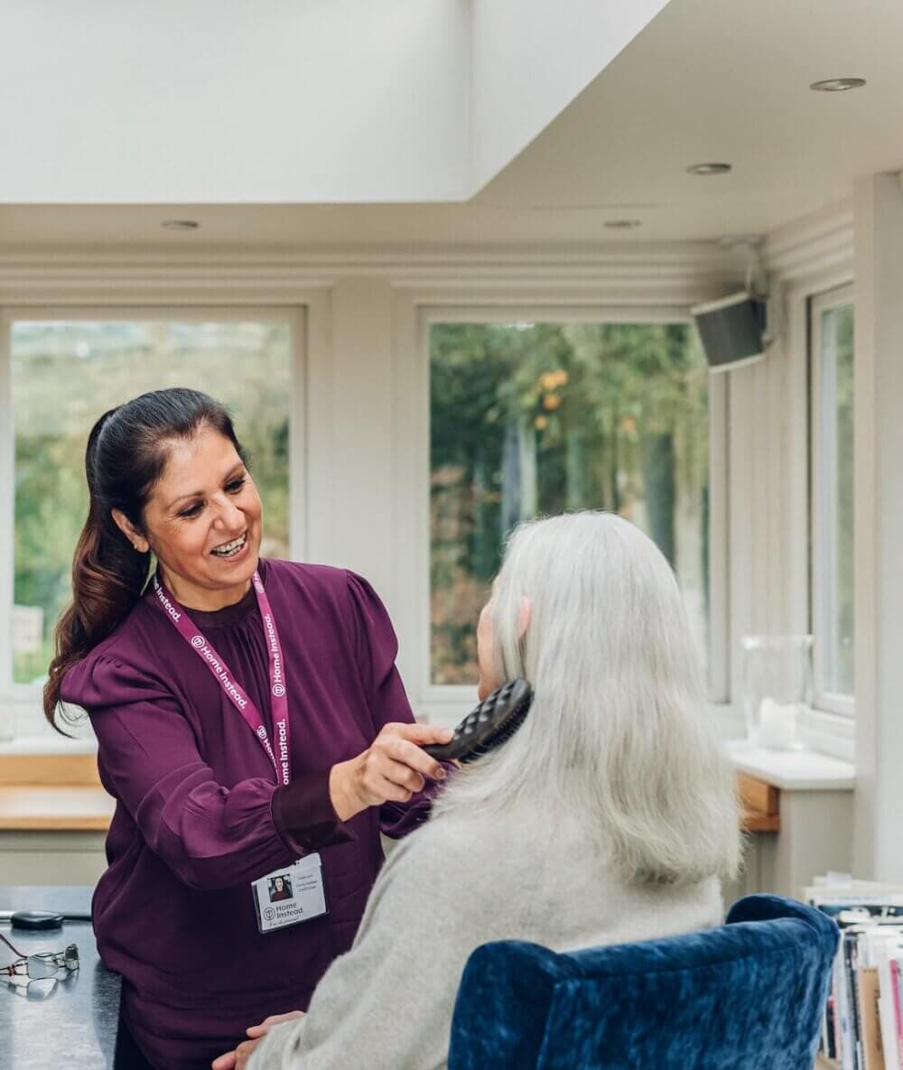A caregiver in a maroon shirt helps brush a senior woman's hair in a bright, sunlit room. - Home Instead