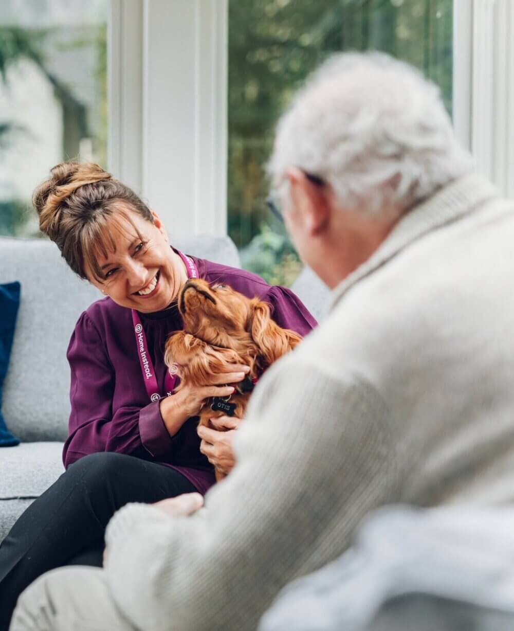 Woman in purple shirt and man with white hair petting a small dog on a sofa, smiling and interacting warmly. - Home Instead