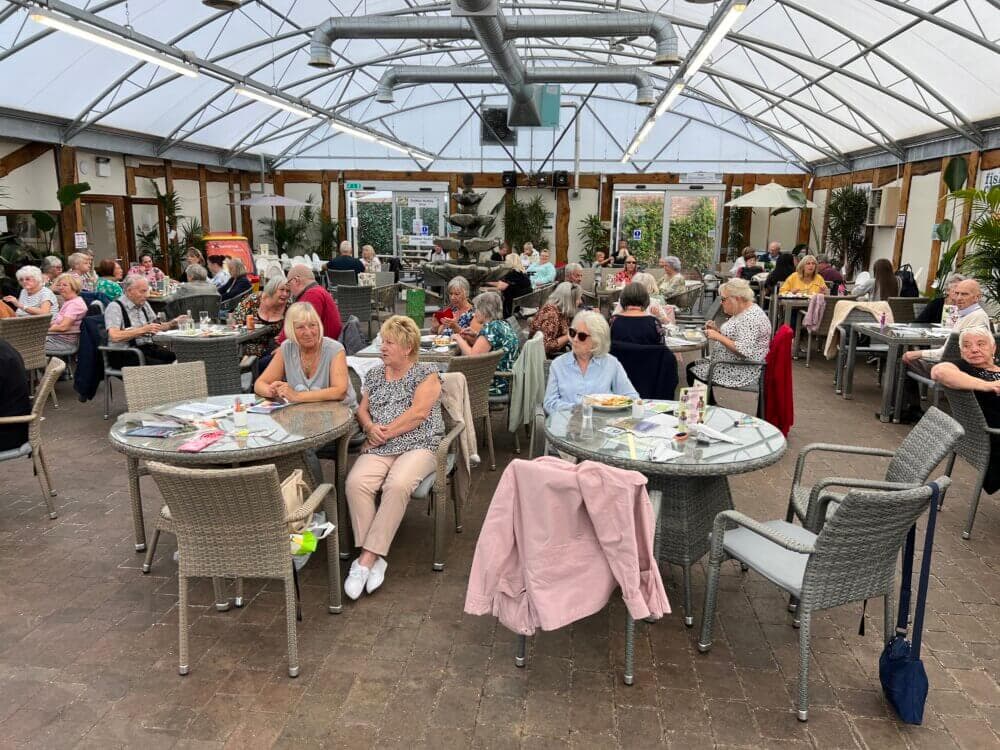 A large group of senior citizens seated at tables inside a greenhouse cafe, having lunch and socializing. - Home Instead