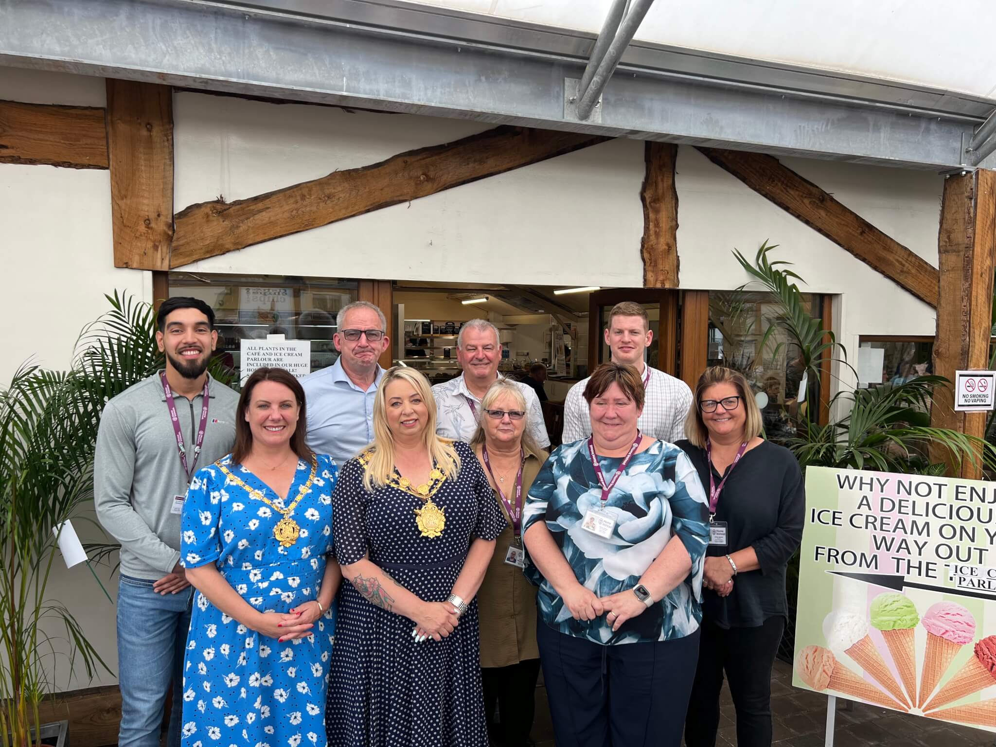 A group of nine people standing together in front of a wooden structure with an ice cream sign nearby. - Home Instead