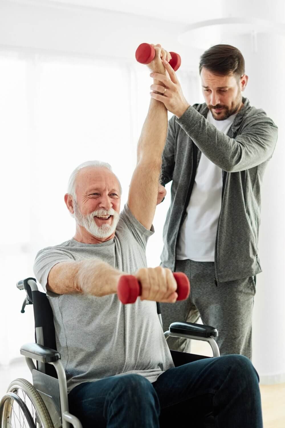A senior man in a wheelchair lifts weights with help from a standing man in a tracksuit. - Home Instead
