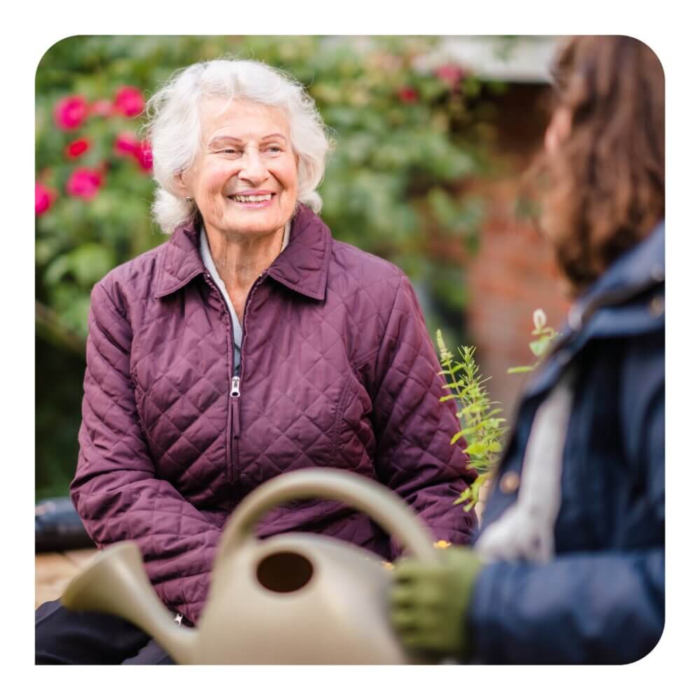 An elderly woman in a purple jacket smiles while talking to a person holding a watering can in a garden. - Home Instead