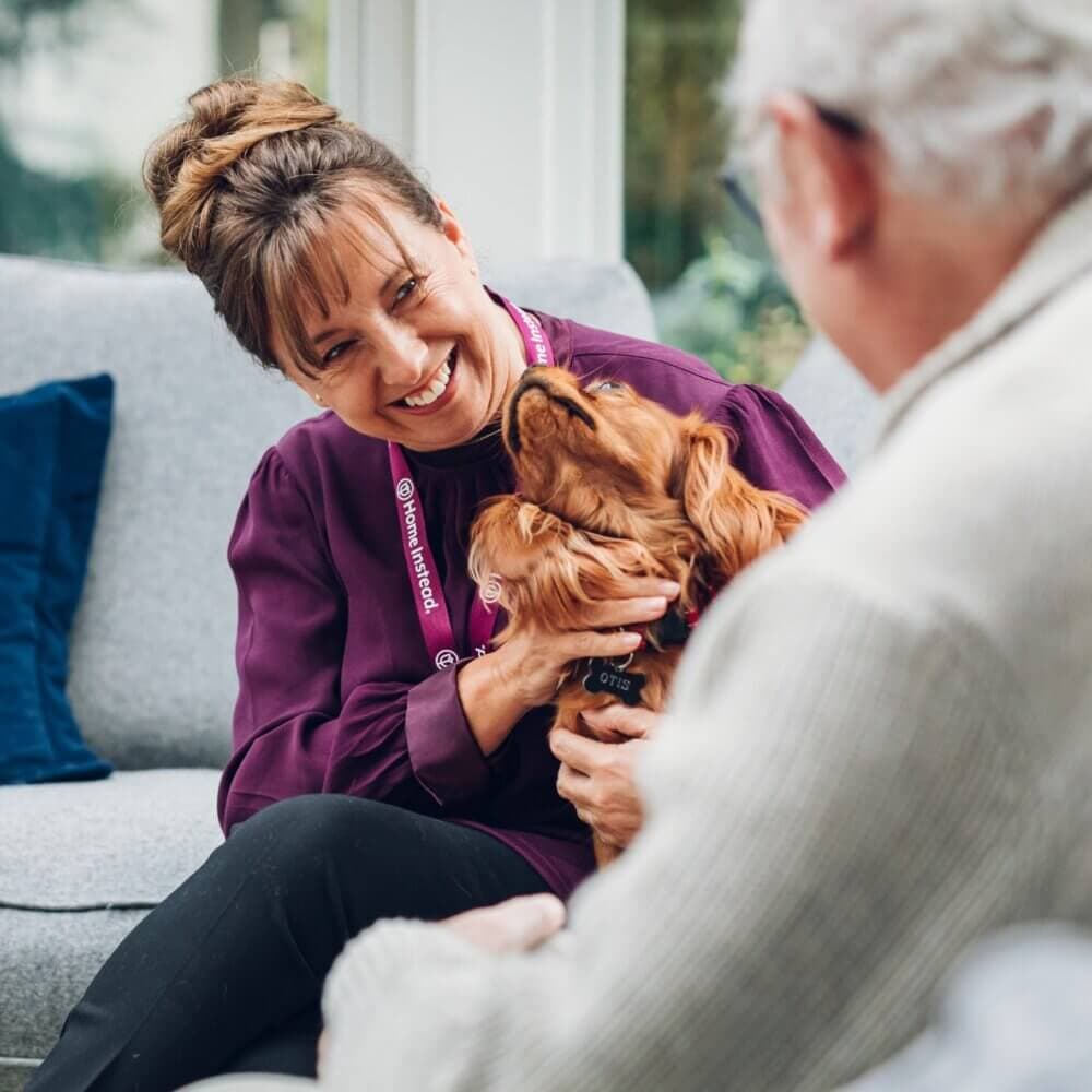 A woman smiles at an elderly man while petting a dog, all sitting on a couch in a cozy room. - Home Instead