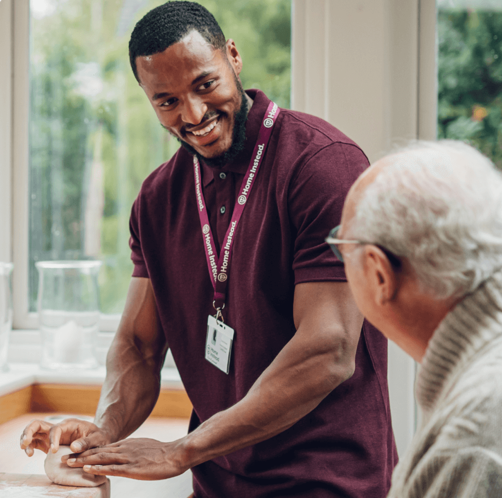 A young male caregiver in a maroon shirt smiling and assisting an elderly man in a bright, homey setting. - Home Instead