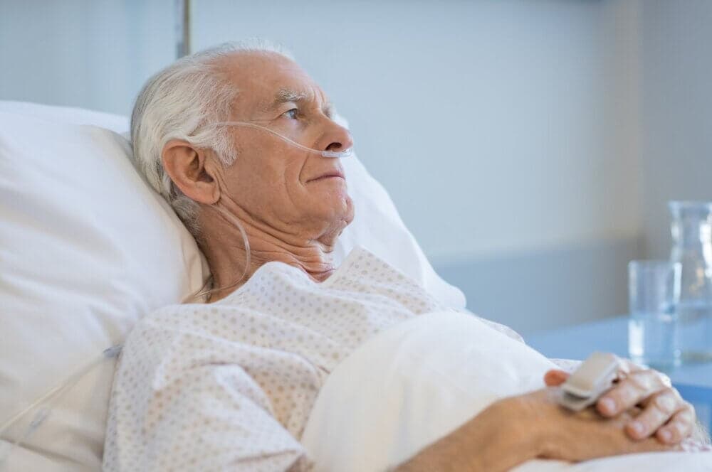 Elderly man with oxygen tube lying in hospital bed, covered in a white blanket, and looking thoughtful. - Home Instead