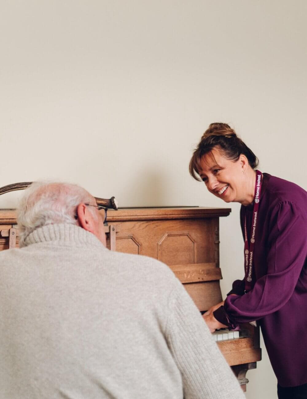 An elderly man in a gray sweater plays the piano while a smiling woman in a purple blouse stands beside him. - Home Instead
