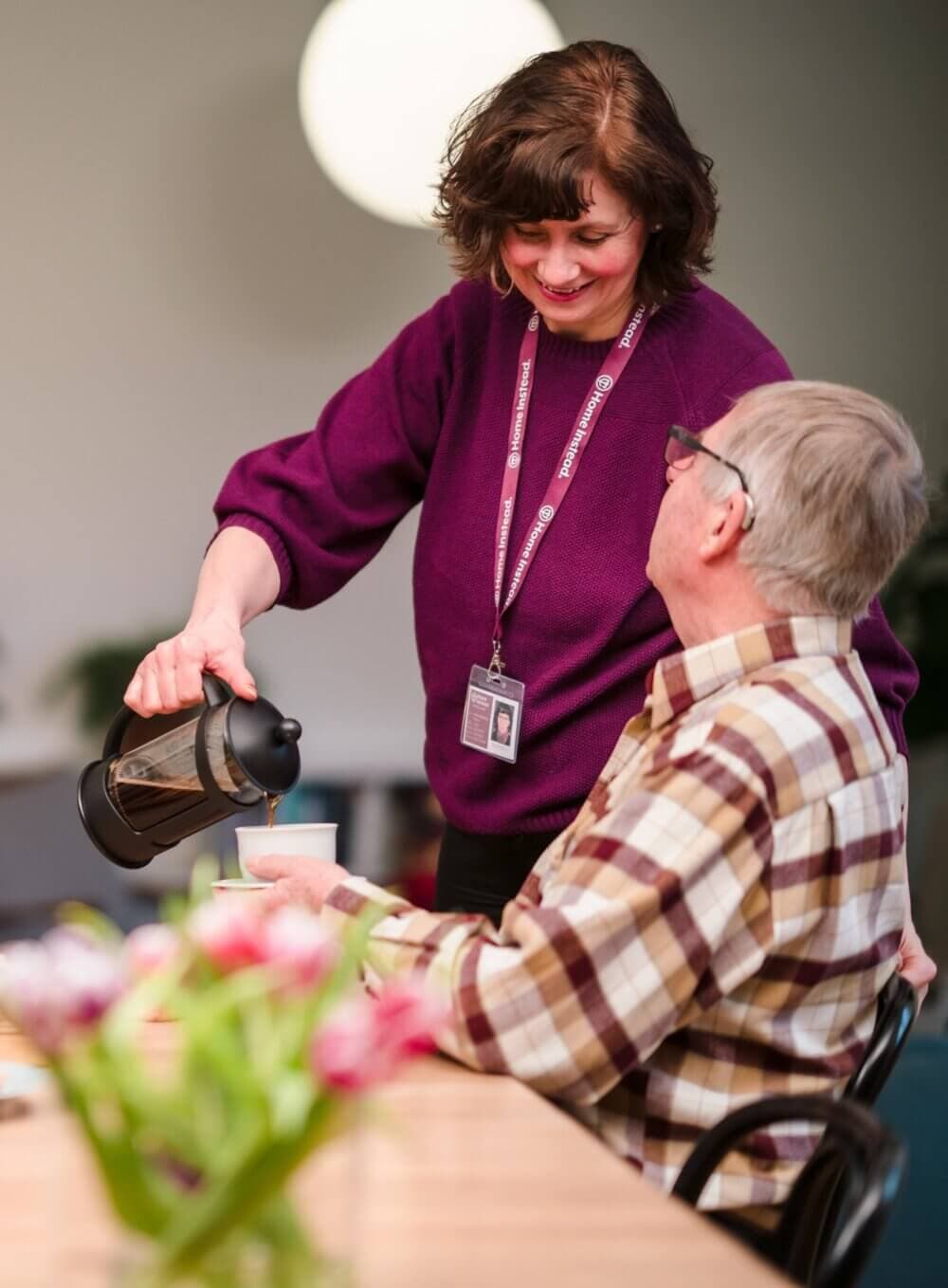 A woman in a purple sweater pours coffee for an elderly man at a table with a bouquet of flowers. - Home Instead