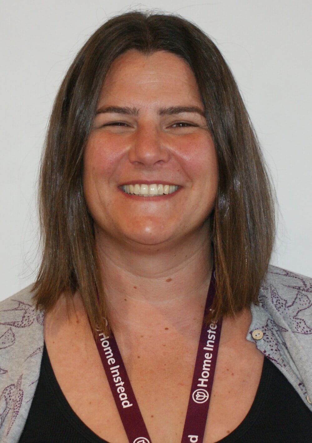 A woman smiling, wearing a Home Instead lanyard, and a light-colored cardigan over a black top. - Home Instead
