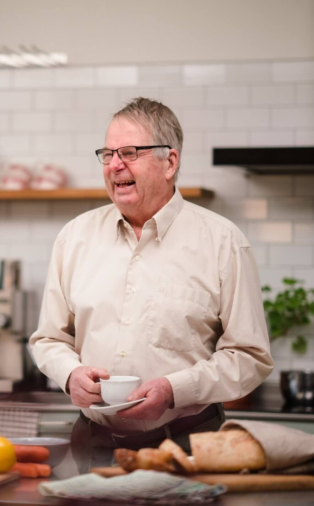 Elderly man in glasses and a beige shirt, smiling and holding a cup in a kitchen with bread and carrots on the counter. - Home Instead