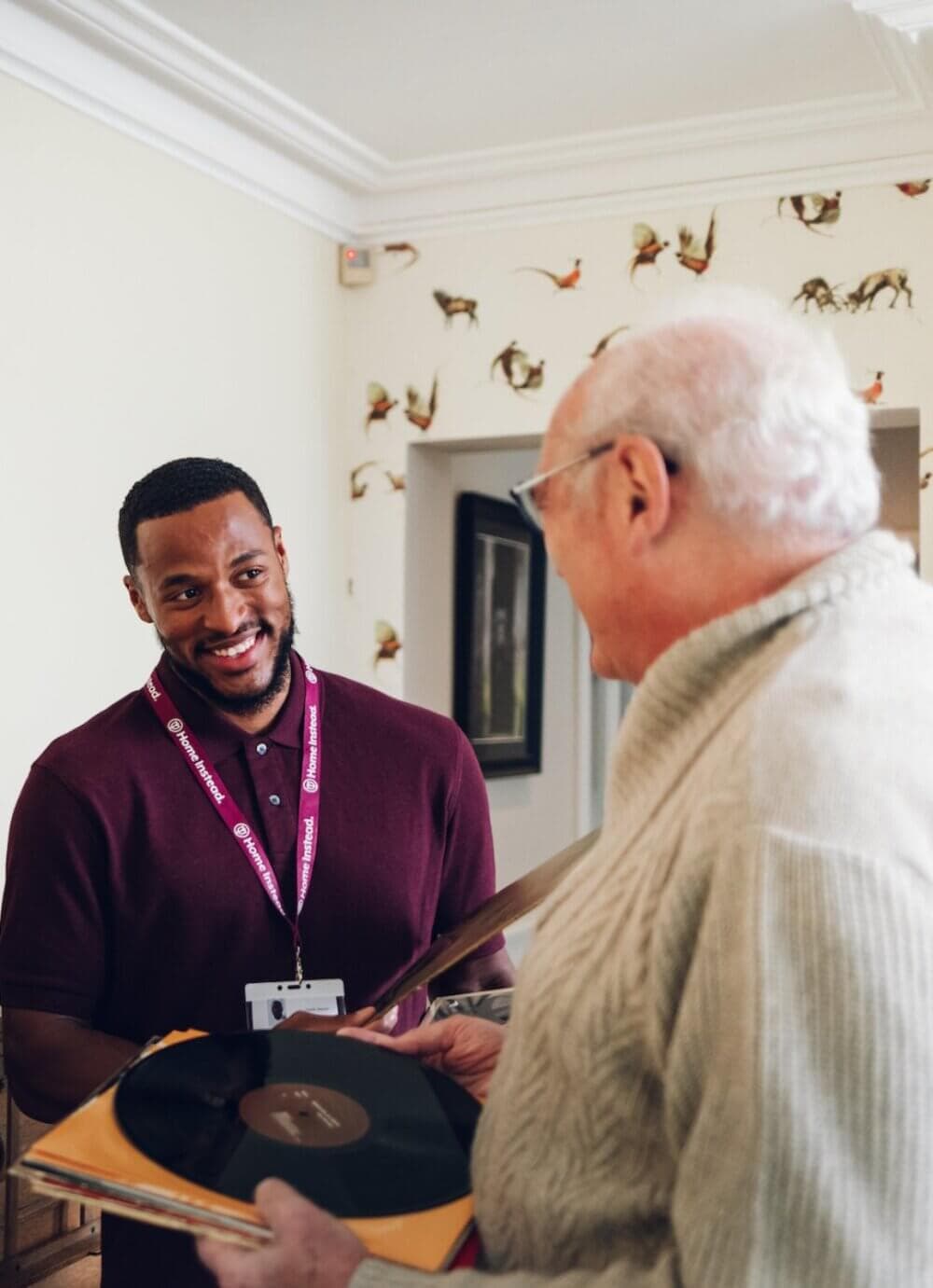 Young man in a maroon shirt smiling at an older man holding a vinyl record in a cozy room with bird wallpaper. - Home Instead