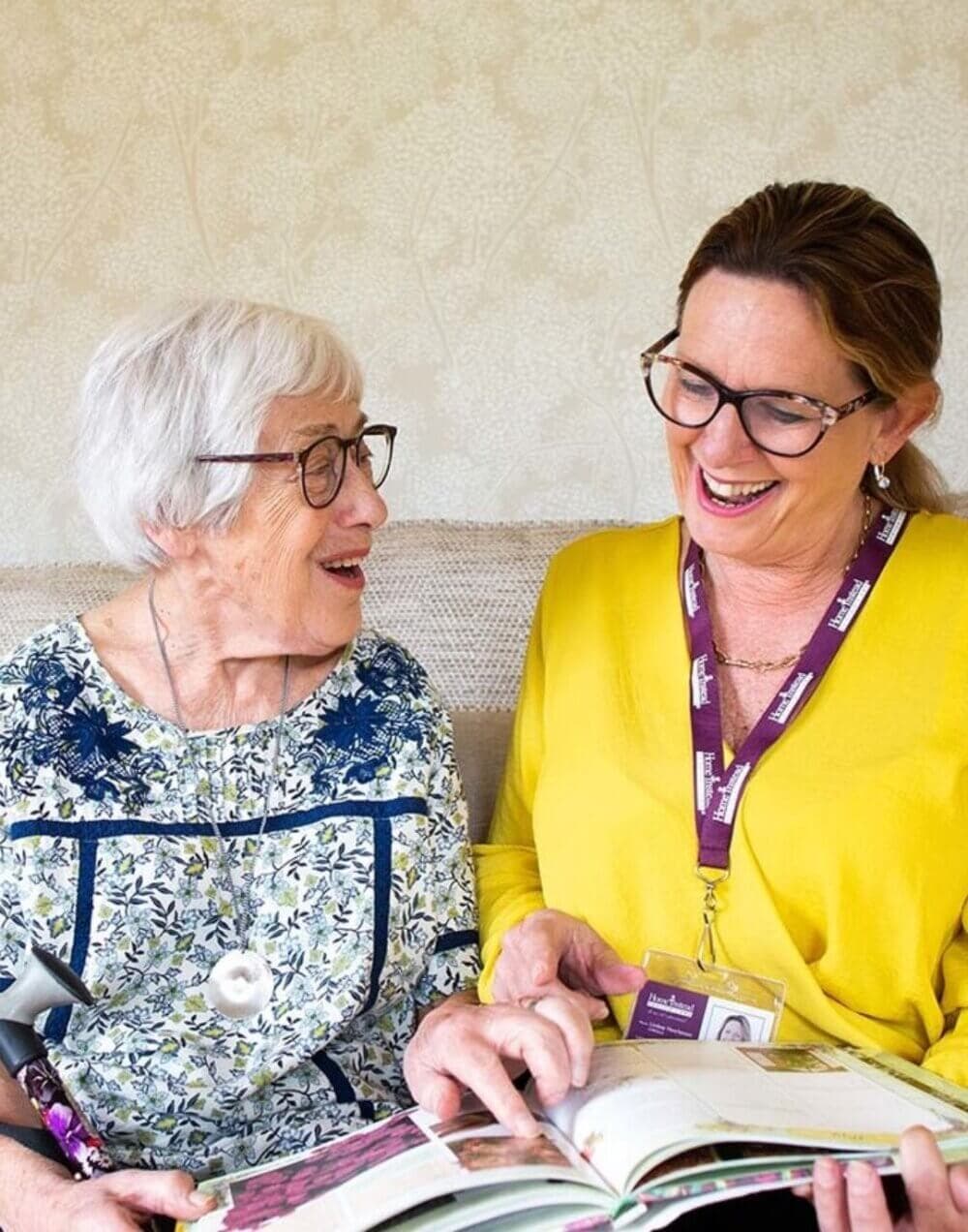 An elderly woman and a younger woman with glasses smile while looking at a book together. - Home Instead