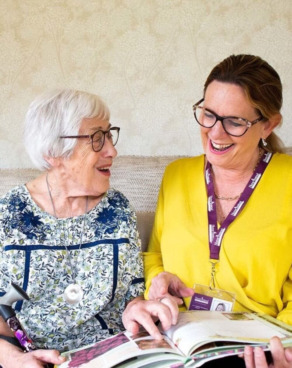 An elderly woman and a caregiver smile while looking at a book together. - Home Instead