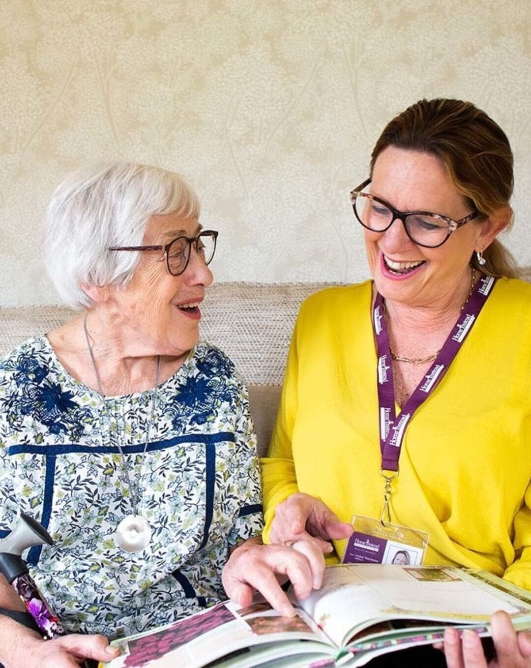 An elderly woman and a caregiver smile while looking at a book together. - Home Instead
