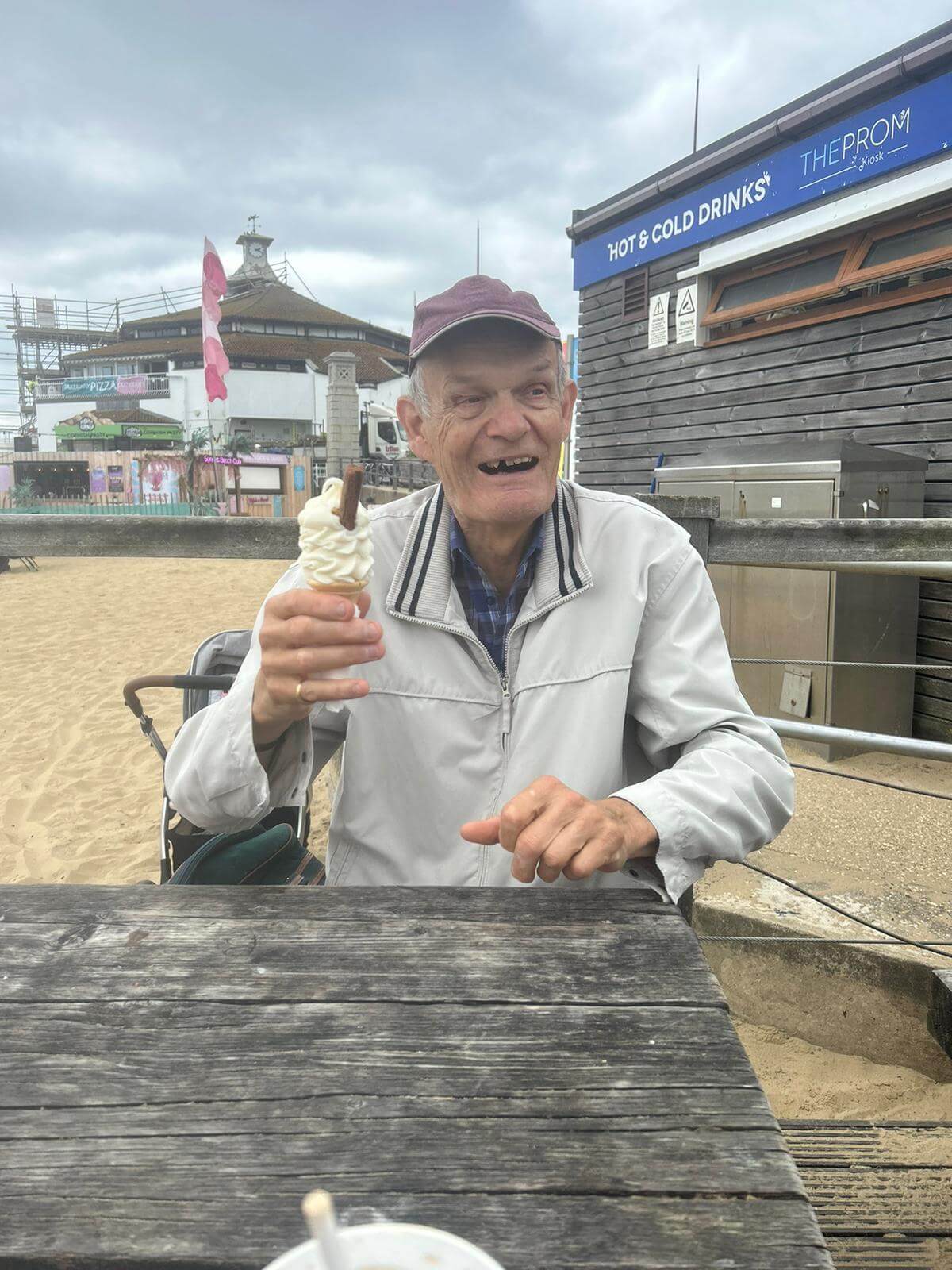 Older man smiling and holding ice cream while sitting at a picnic table near a beach and amusement park. - Home Instead