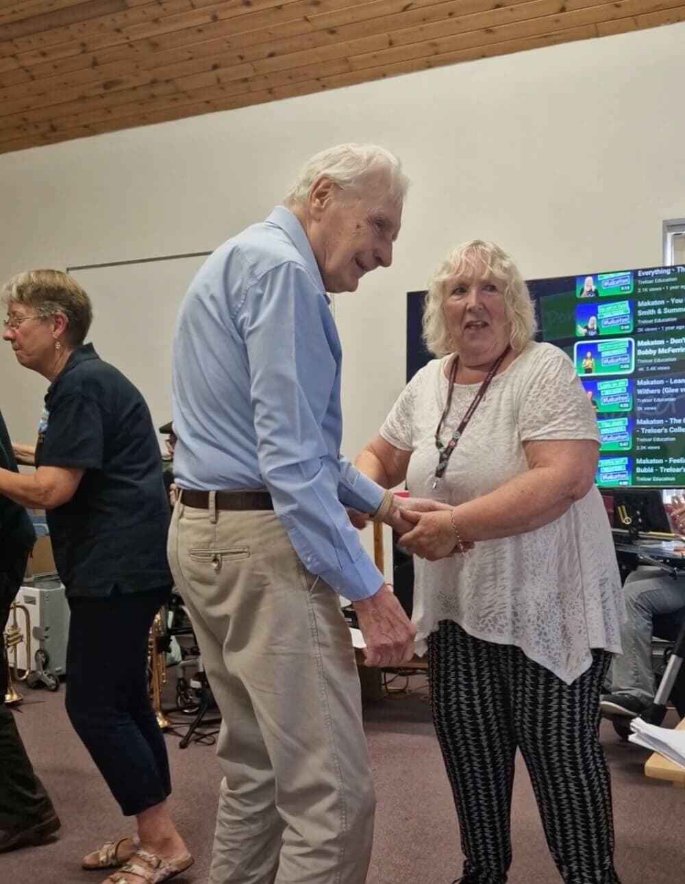 An elderly man and woman smile while holding hands in a room; another woman stands in the background. - Home Instead