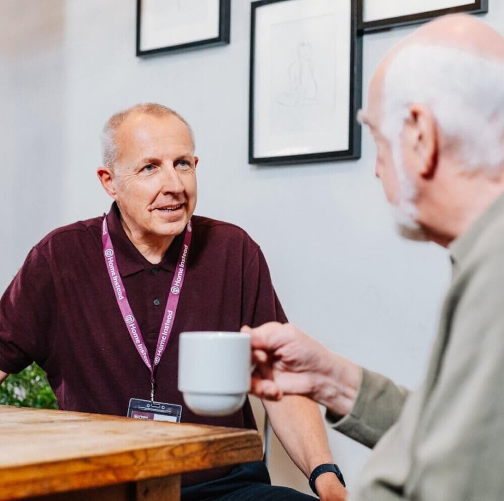 Two men sitting and conversing while holding mugs in a casual setting with framed artwork on the wall behind them. - Home Instead