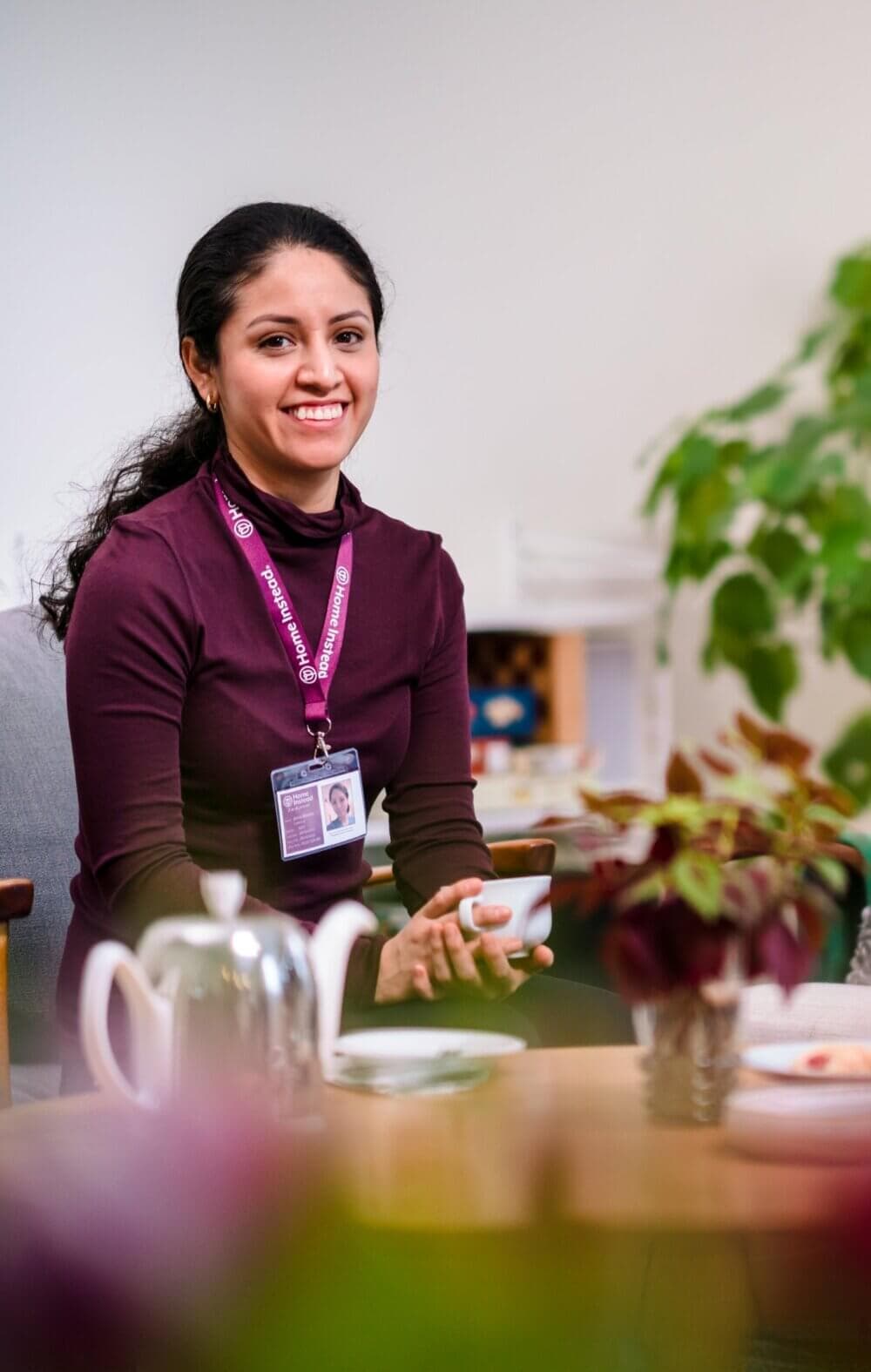 Smiling woman with lanyard and ID badge, holding a cup, sitting at a table with a teapot, cup, and plants. - Home Instead